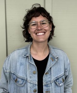 Rachel wearing glasses, a jean jacket, and standing in front of a tan wall.