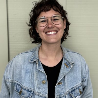 Rachel wearing glasses, a jean jacket, and standing in front of a tan wall.