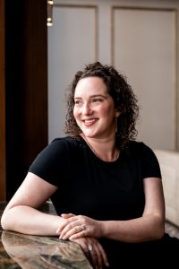 Sophia seated looking to her right with hands folded wearing a black shirt.