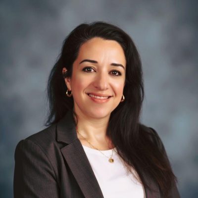 Sheila sitting in front of a grey screen wearing a charcoal blazer and white shirt.