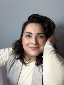 Alma in front of a grey screen wearing a white shirt and grey vest rested her head on her hand