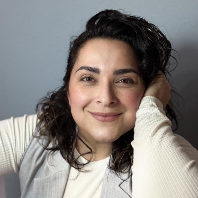 Alma in front of a grey screen wearing a white shirt and grey vest rested her head on her hand